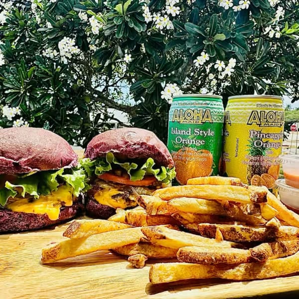Two Taro Hut burgers with fries and Aloha Maid drinks on a wooden board under a flowering tree.