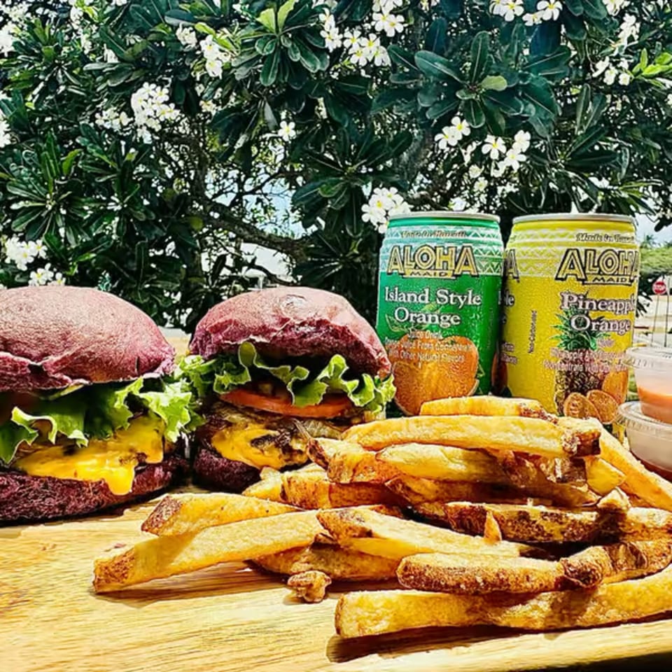 Two Taro Hut burgers with fries and Aloha Maid drinks on a wooden board under a flowering tree.