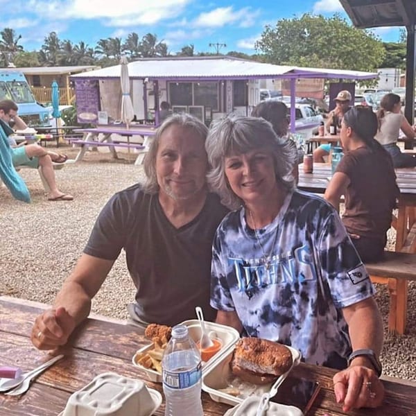 A couple smiles at a picnic table with Taro Hut burgers and fries, surrounded by other diners.