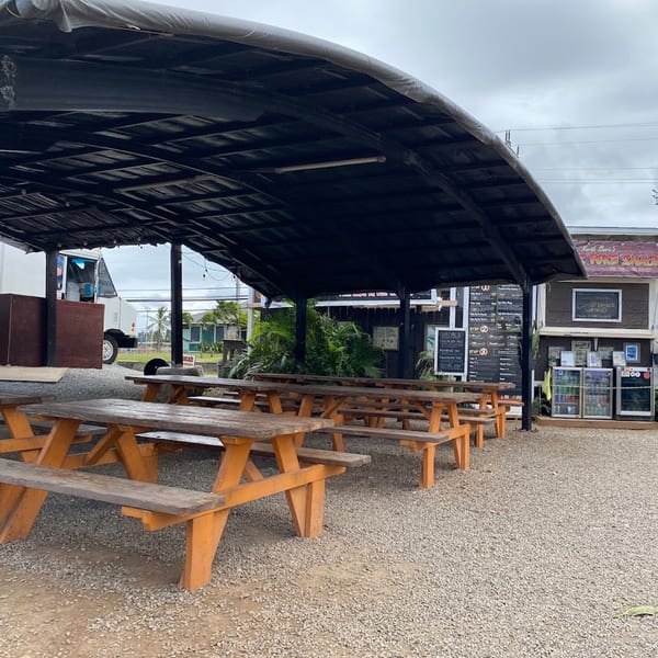 Outdoor seating area with multiple wooden picnic tables under a large black awning at Taro Hut.