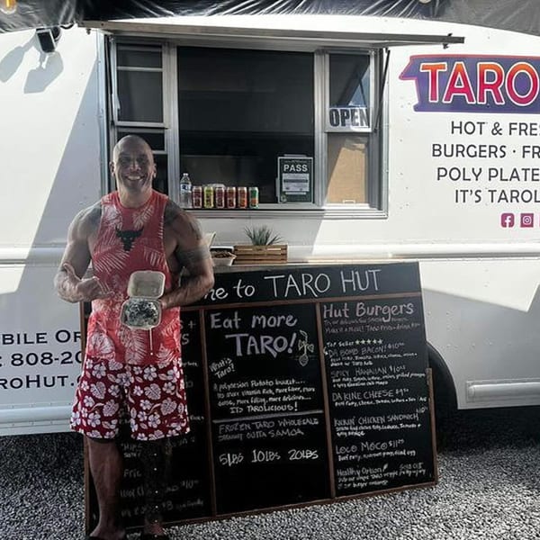 A man in a red tank top and shorts smiles, holding a burger container in front of the Taro Hut food truck.