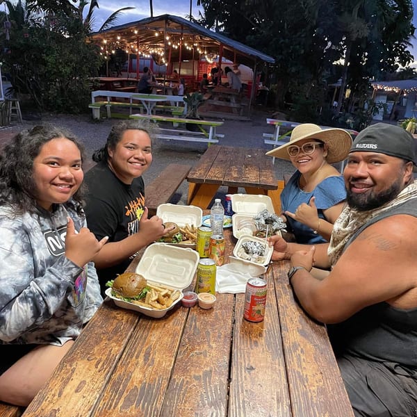 Four happy customers enjoying Taro Hut burgers and drinks at an outdoor picnic table at dusk.