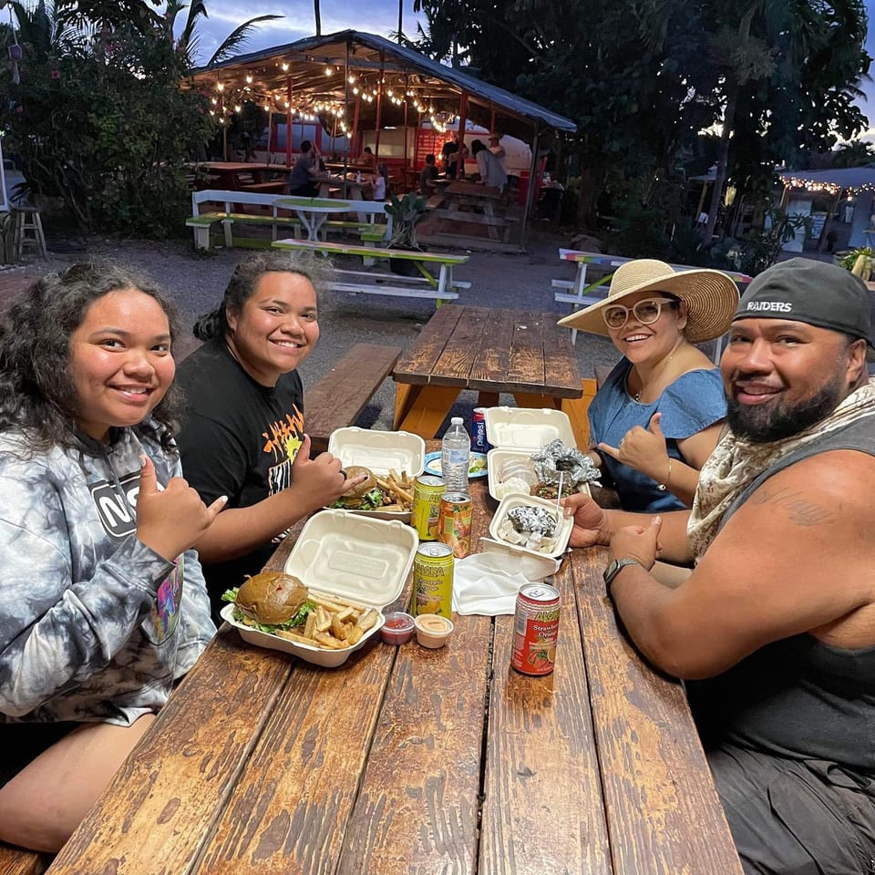 Four happy customers enjoying Taro Hut burgers and drinks at an outdoor picnic table at dusk.