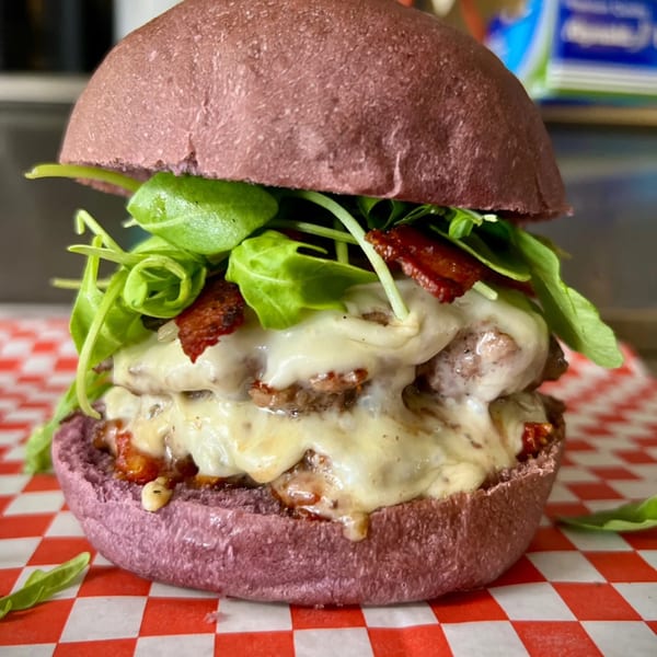 A Taro Hut burger with a purple bun, double patty, melted cheese, bacon, and arugula on checkered paper.