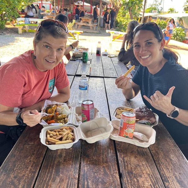 Two women smiling at a wooden picnic table with Taro Hut burgers, fries, and drinks.