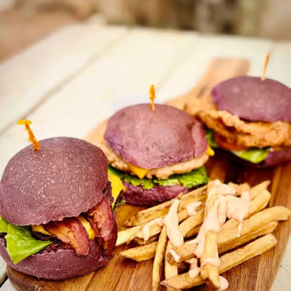 Three Taro Hut purple-bun burgers with fries on a wooden board, showcasing unique burger creations.