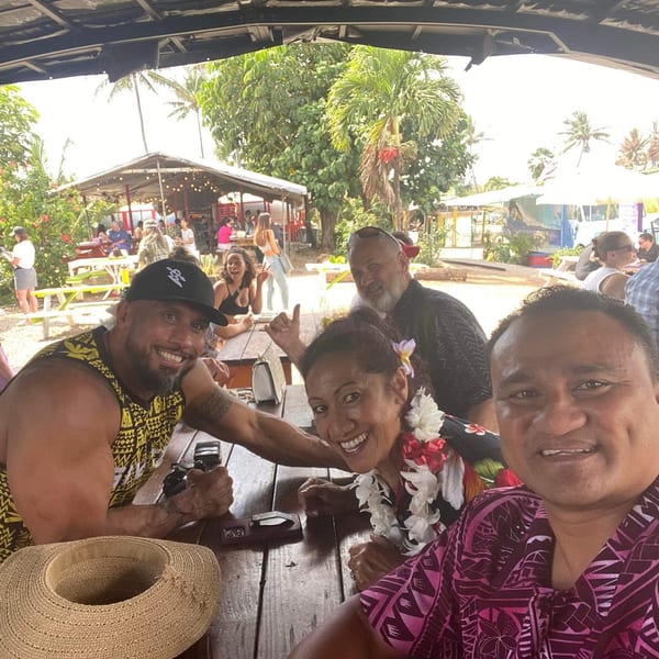 Four smiling people pose for a selfie at an outdoor restaurant with palm trees and food trucks.
