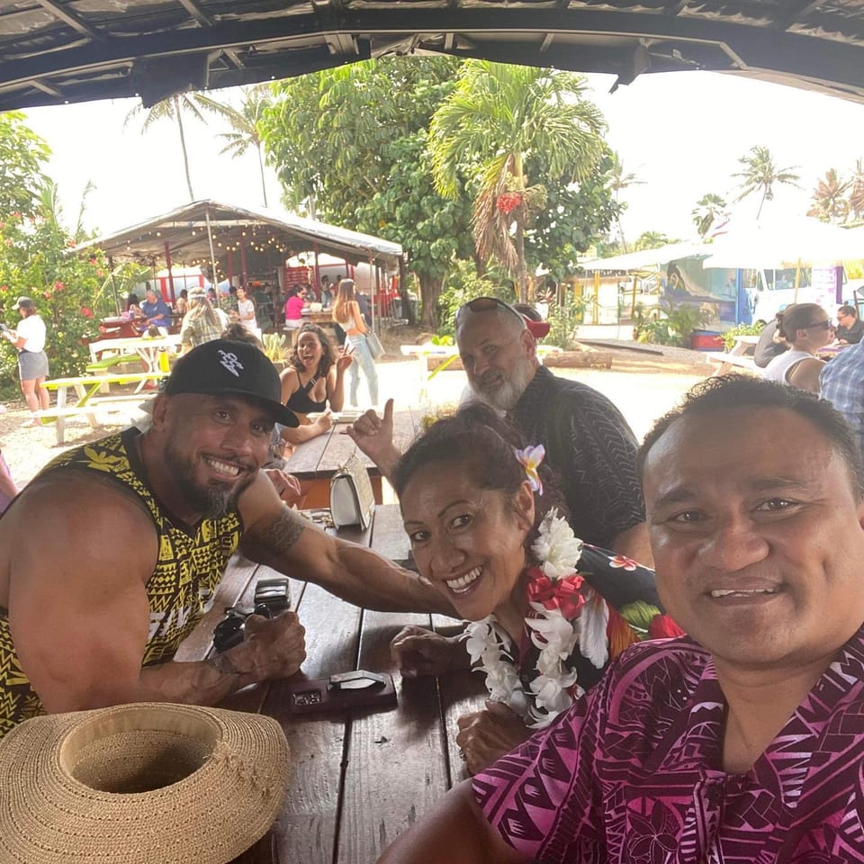 Four smiling people pose for a selfie at an outdoor restaurant with palm trees and food trucks.