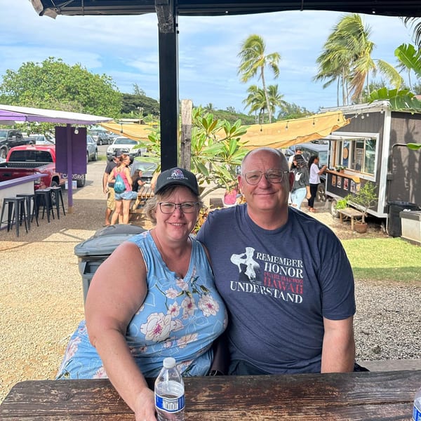 A smiling couple sits at a picnic table at Taro Hut, with food trucks and palm trees in the background.